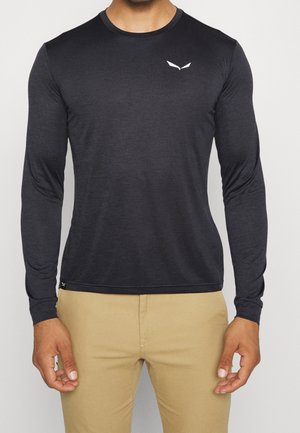 Mężczyzna ubrany w dopasowaną czarną koszulkę z długim rękawem z małym białym logo na piersi oraz beżowe spodnie, stojący z rozluźnionymi ramionami.