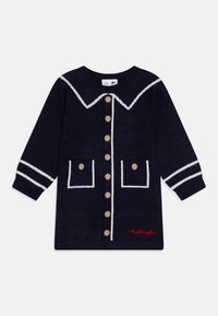 Marineblå strikket kjole med hvit krage og stripete mansjetter. Har dekorative knapper og to frontlommer med beige prikker. Rødt "Paddington" broderi.