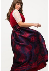 Frau in rotem Mieder und weißen Spitzenärmeln, die einen marineblauen Rock mit roten floralen Mustern auf einem weißen Hintergrund dreht.