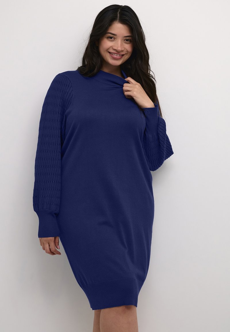 Abito lavorato a maglia in blu scuro con colletto alto, caratterizzato da maniche a palloncino testurizzate. Termina sopra il ginocchio con polsini a coste per una vestibilità rilassata.
