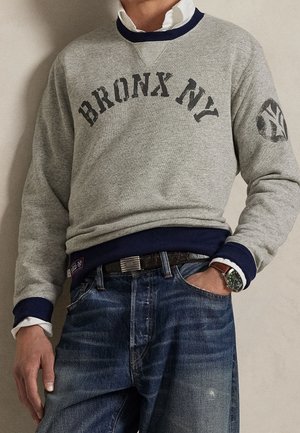 Mężczyzna w szarej bluzie z napisem "BRONX NY", pod spodem biała koszula, niebieskie dżinsy, czarny pasek i zielony zegarek, ręka w kieszeni.