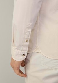 Camisa de botones beige claro con mangas largas, que presenta un dobladillo redondeado, costuras visibles y un detalle de botón en el puño.