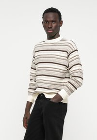 Pull en tricot rayé crème avec des accents marron, doté de poignets et d'un col côtelés. Conçu avec une coupe décontractée et des motifs texturés.