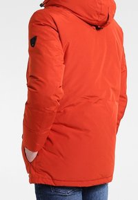 Červená izolovaná bunda s kapucí, na rameni s nášivkou s logem a elastickými manžetami. Materiál působí hladce a odolně vůči povětrnostním vlivům.