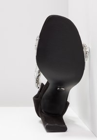 Chaussure noire à talon haut avec un bout carré et des brides décoratives argentées, vue de la semelle inférieure sur fond blanc.