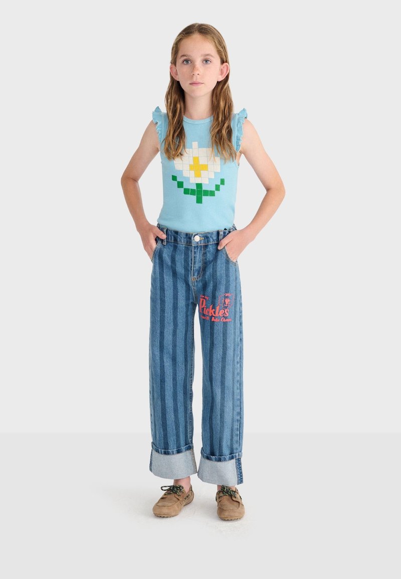 Pige står med hænderne i lommerne iført en blå, ærmeløs top med pixeleret blomst, stribede, vide jeans og beige sko.