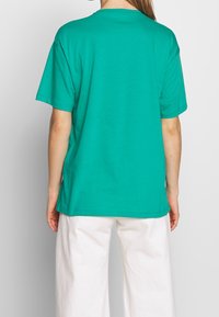 T-shirt en coton teal avec des manches courtes et un col rond, affiché de dos. Associé à un pantalon blanc ample.