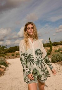 Lichtgewicht, doorschijnend wit shirt met groene palmblaadjesprints, lange mouwen en een knoopsluiting aan de voorkant. De zoom heeft een gemusterde accent.