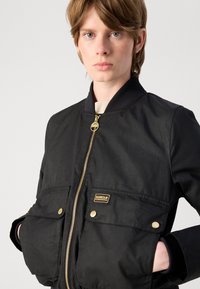 Μαύρο bomber jacket με χρυσές λεπτομέρειες, φερμουάρ μπροστά, ριμπ λαιμό και δύο μπροστινές τσέπες. Περιλαμβάνει ένα επώνυμο λογότυπο στην αριστερή τσέπη.