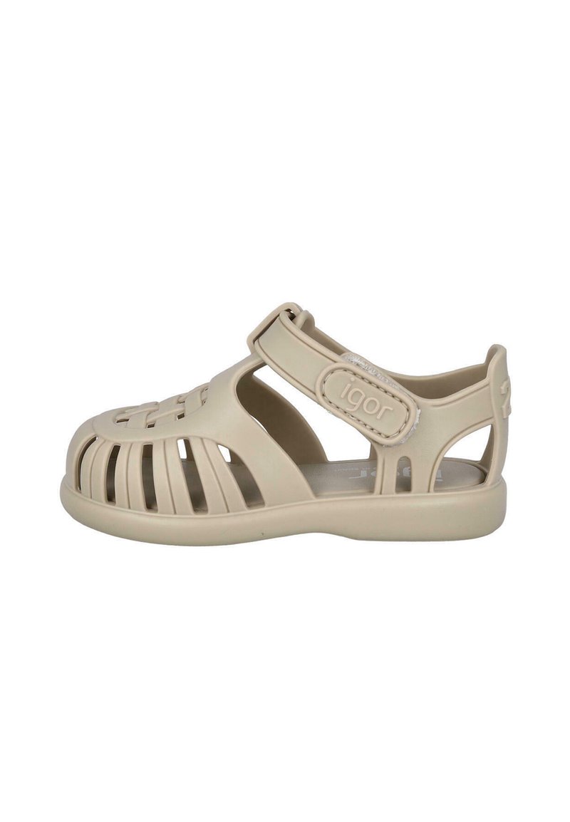 Sandalias de plástico beige con parte superior abierta que presentan un diseño ranurado, correa ajustable y punta redondeada. Construcción ligera para facilitar su uso.