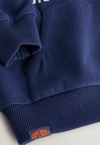 Sudadera azul marino con puños y dobladillo de canalé, que presenta una tela texturizada. Incluye una etiqueta naranja "SUPERDRY" en la parte inferior izquierda.
