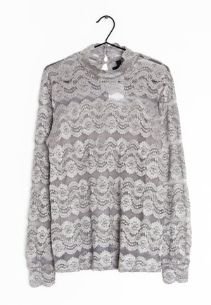 Top lungo a maniche, in pizzo grigio con motivi floreali, caratterizzato da un colletto alto e una texture trasparente, appeso a un gancio nero su uno sfondo bianco.