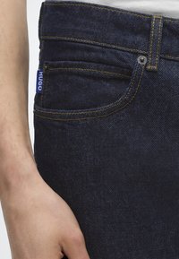 Venstre hånd hviler ved siden af mørkeblå denimjeans med gul syning og et lille blåt "HUGO" mærke på lommen.