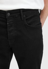 Jeans de mezclilla negra con un diseño clásico de cinco bolsillos, que presentan un cierre de botón en la parte delantera y detalles de costura sutiles en los bolsillos.