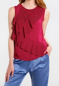 Bezramínkový top v hluboké burgundské barvě s diagonálním skládáním a volánem přes přední stranu; hladká tkanina; doplněný lesklými modrými saténovými kalhotami.