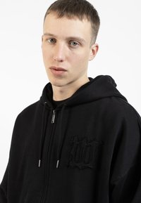 Svart hettejakke med glidelås laget av myk stoff, med lomme foran og en teksturert brodert design på brystet. Glatt glidelåsmekanisme.