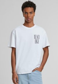 T-shirt en coton blanc à manches courtes et encolure ras du cou. Présente du texte vertical noir "REACH HIGH" sur le côté gauche. Coupe décontractée.