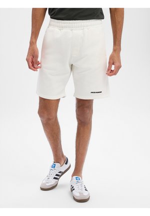 Man draagt witte shorts met elastische taille en 'PEGADOR'-logo en witte Adidas sneakers met zwarte strepen, lopend tegen een effen achtergrond.