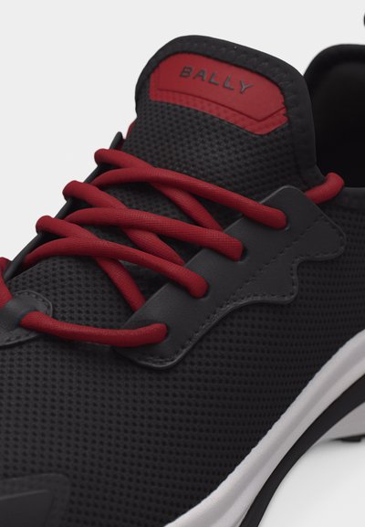 Baskets Bally noires avec lacets rouges, étiquette rouge sur la languette, et semelle blanche, montrant des coutures détaillées et un matériau texturé.