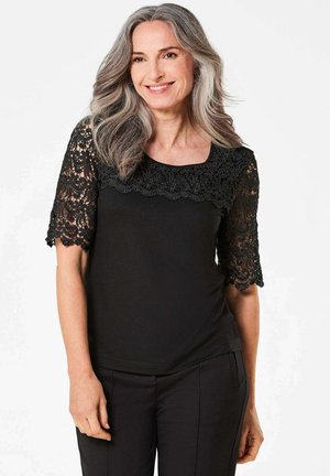 Schwarzes kurzärmeliges Oberteil mit einem Spitzenmuster und floralem Design an den Ärmeln, kombiniert mit schwarzen maßgeschneiderten Hosen.
