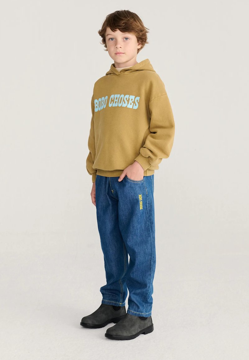 Garçon debout portant un sweat à capuche marron avec texte bleu, un jean bleu avec écriture jaune et des bottines noires sur un fond uni.