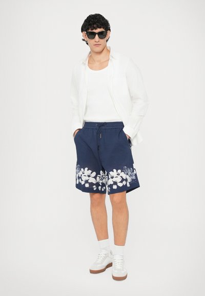 Shorts en coton bleu marine avec motif floral blanc, taille élastique, poches latérales, assortis à un débardeur blanc et une chemise blanche légère.