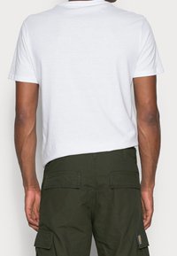 Man in een eenvoudige witte korte mouwen t-shirt en donker groene cargobroek, gezien van achteren tegen een neutrale achtergrond.