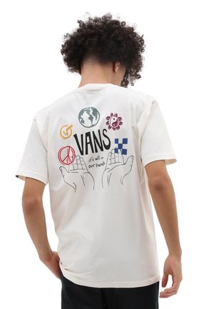 Personne aux cheveux bouclés portant un t-shirt blanc montrant des mains, le signe de la paix, un globe, une fleur et le logo Vans avec le texte « tout est entre nos mains ».