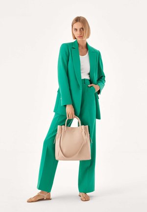 Blazer et pantalons ajustés verts associés à un haut blanc. Tenant un sac fourre-tout en cuir beige avec des poignées doubles et une texture subtile. Sandales plates.