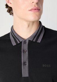 Svart polo skjorta med en grårandig krage, tre knappar och en diskret präglad "BOSS"-logotyp på bröstet. Mjuk, strukturerad tyg.