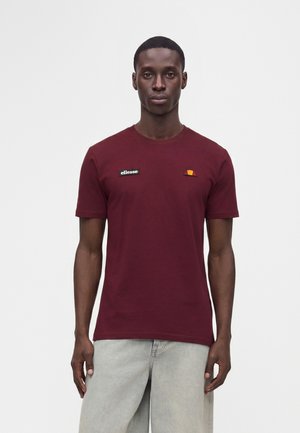 Uomo che indossa una t-shirt aderente color bordeaux con toppe del logo ellesse sul petto e jeans grigio chiaro, in piedi davanti a uno sfondo neutro.