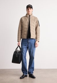 Beige vadderad jacka med hög krage, svarta detaljer och dragkedja framtill, tillsammans med blåjeans och svarta skor, hållande en svart väska.