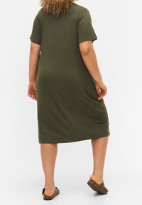 Olivově zelené žebrované šaty s krátkými rukávy, délka do poloviny lýtek, boční kapsy a volný střih. Modelka má na sobě hnědé nazouvací boty.