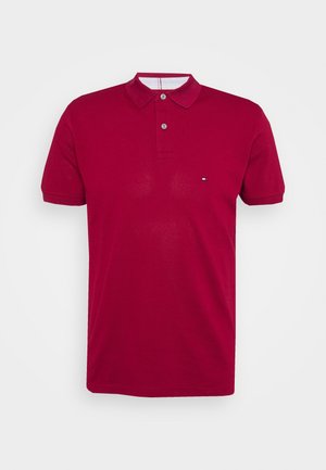 Polo rouge à manches courtes avec deux boutons, col côtelé et petit logo sur le côté gauche de la poitrine sur fond blanc.