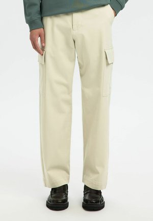 Pantalon cargo beige avec poches latérales, porté avec des chaussures noires et un haut vert à manches longues, vue de la taille aux pieds.