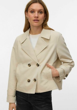 Kvinde iført en beige dobbeltradet blazer med fire skildpaddeskal-knapper, den ene hånd i lommen, kombineret med sorte bukser og en hvid top.