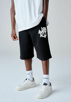 Inimene, kes kannab musta poolpikka pükst, millel on valge "AllSaints London" logo, valgeid sokke, valgeid tossusid ja lahtist valget t-särki.