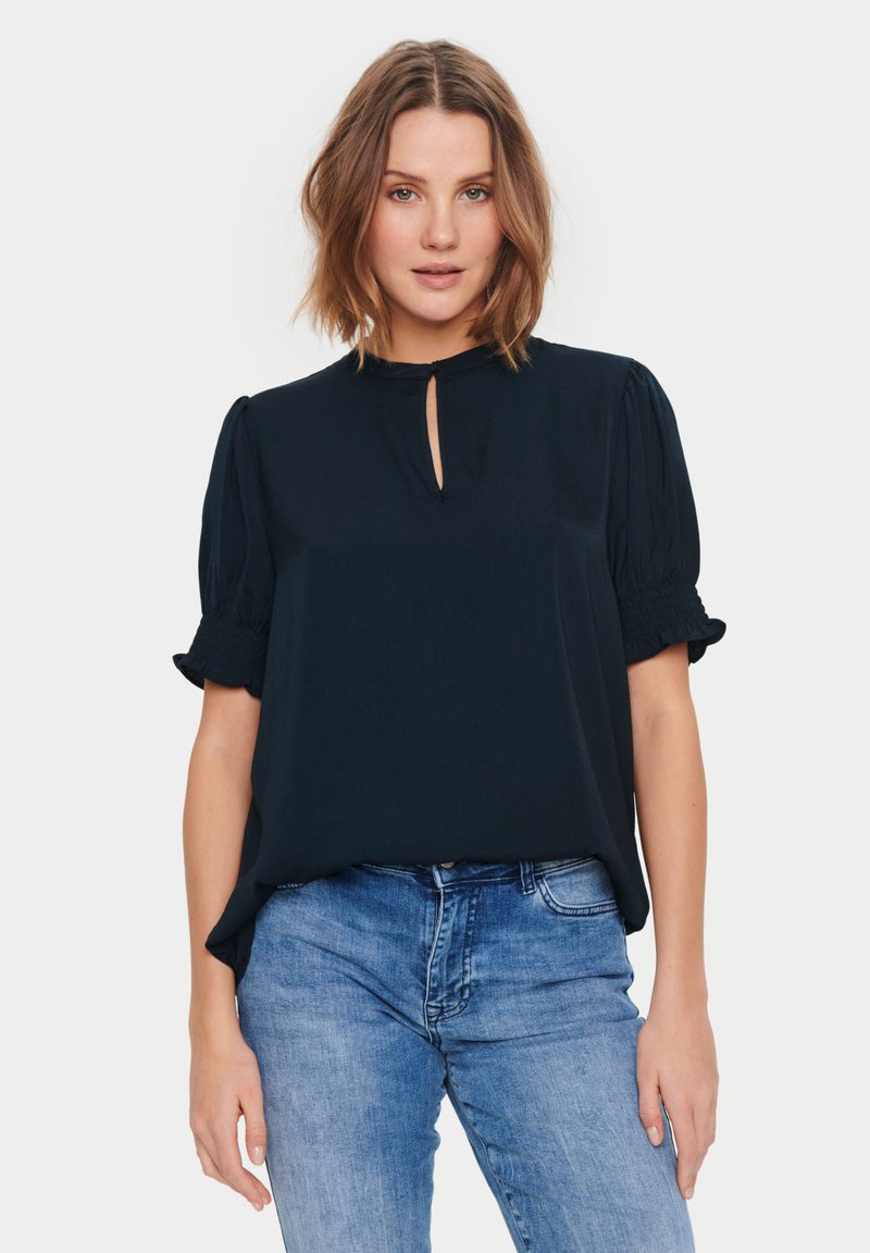 Blusa blu navy con maniche a palloncino corte e scollatura a chiave, realizzata in un tessuto morbido, abbinata a jeans in denim azzurro chiaro.