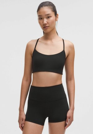 Young woman with straight dark hair tied back wearing black cropped tank top and black high-waisted fitted shorts against plain background.