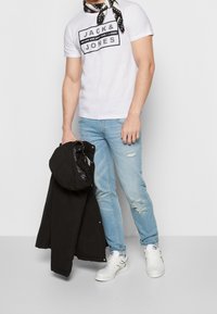 White t-shirt with black graphic text, light blue ripped jeans, and a black jacket held in one hand. Wearing white sneakers and a black scarf.