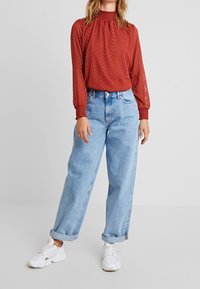 Backstensröd, strukturerad blus med smockade manschetter, i kombination med löst sittande ljusblå jeans och vita sportskor.