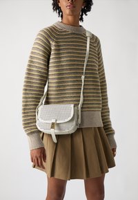 Borsa a tracolla in pelle bianca perforata con una chiusura dorata, indossata sopra un maglione a righe e una gonna plissettata marrone.