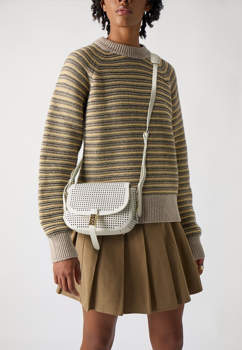 Borsa a tracolla in pelle bianca perforata con una chiusura dorata, indossata sopra un maglione a righe e una gonna plissettata marrone.