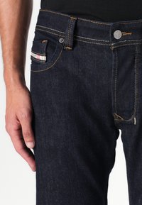 Jeans oscuros de mezclilla, con costuras amarillas, un bolsillo frontal con una etiqueta y un cierre de botón metálico.