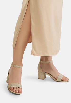 Sandali con tacco glitter dorato, design open toe, cinturini alla caviglia e tacco a blocco, abbinati a un vestito di satin beige con spacco laterale.