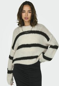 Oversized gebreid sweater in crèmekleur met dikke zwarte strepen. Losse pasvorm met brede mouwen en een gestructureerde afwerking. Gepaard met een zwarte rok.