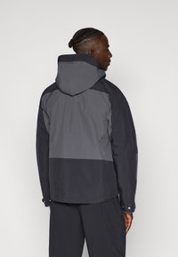 Chaqueta negra y gris con capucha, que presenta un diseño a bloques de color, textura suave y puños ajustables. Estilo minimalista sin logotipos visibles.