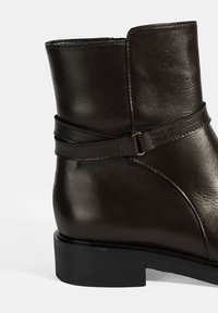 Botte cheville en cuir marron avec une surface lisse, présentant un bout rond élégant, une sangle décorative et un talon bas empilé.