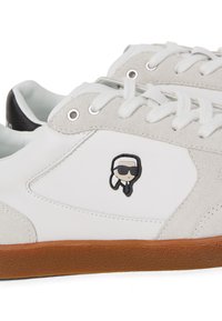 Valged nahast spordijalatsid hallide sametist detailidega, ümmargune must logo päikeseprillidega. Valged paelad ja pruuni kummitald.