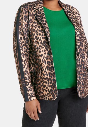 Persoon draagt een blazer met luipaardprint, zwarte streep en tekst op de mouw, groene top en zwarte broek, staand tegen een effen achtergrond.